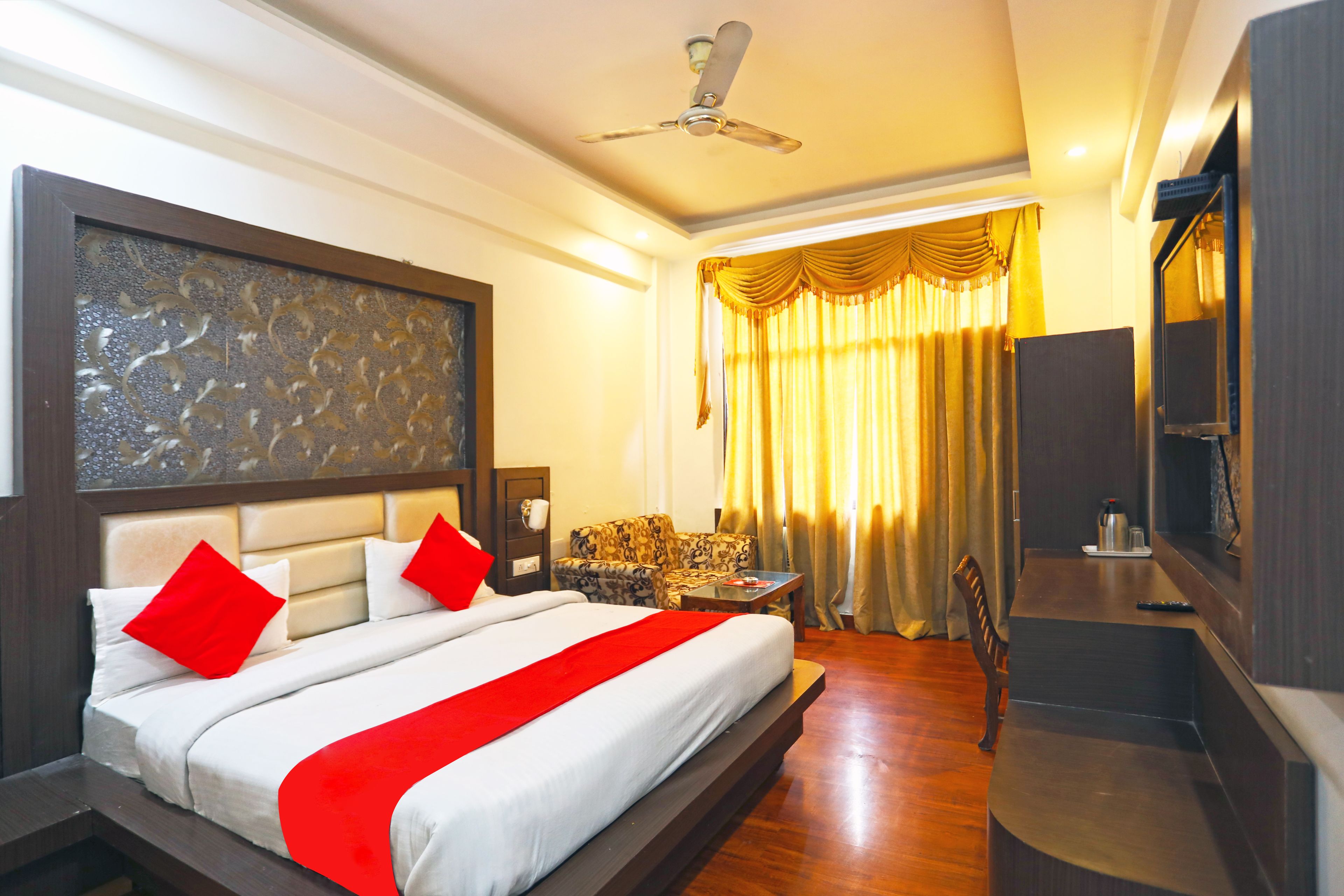 Quarto de hotel com uma cama grande, uma escrivaninha, uma cadeira, uma televisão e um ventilador de teto.