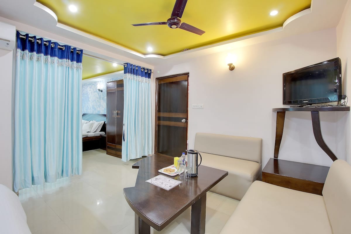 A hotel room with a bed, a dining table with a plate of food, a coffee pot, a TV on a stand, and a ceiling fan.