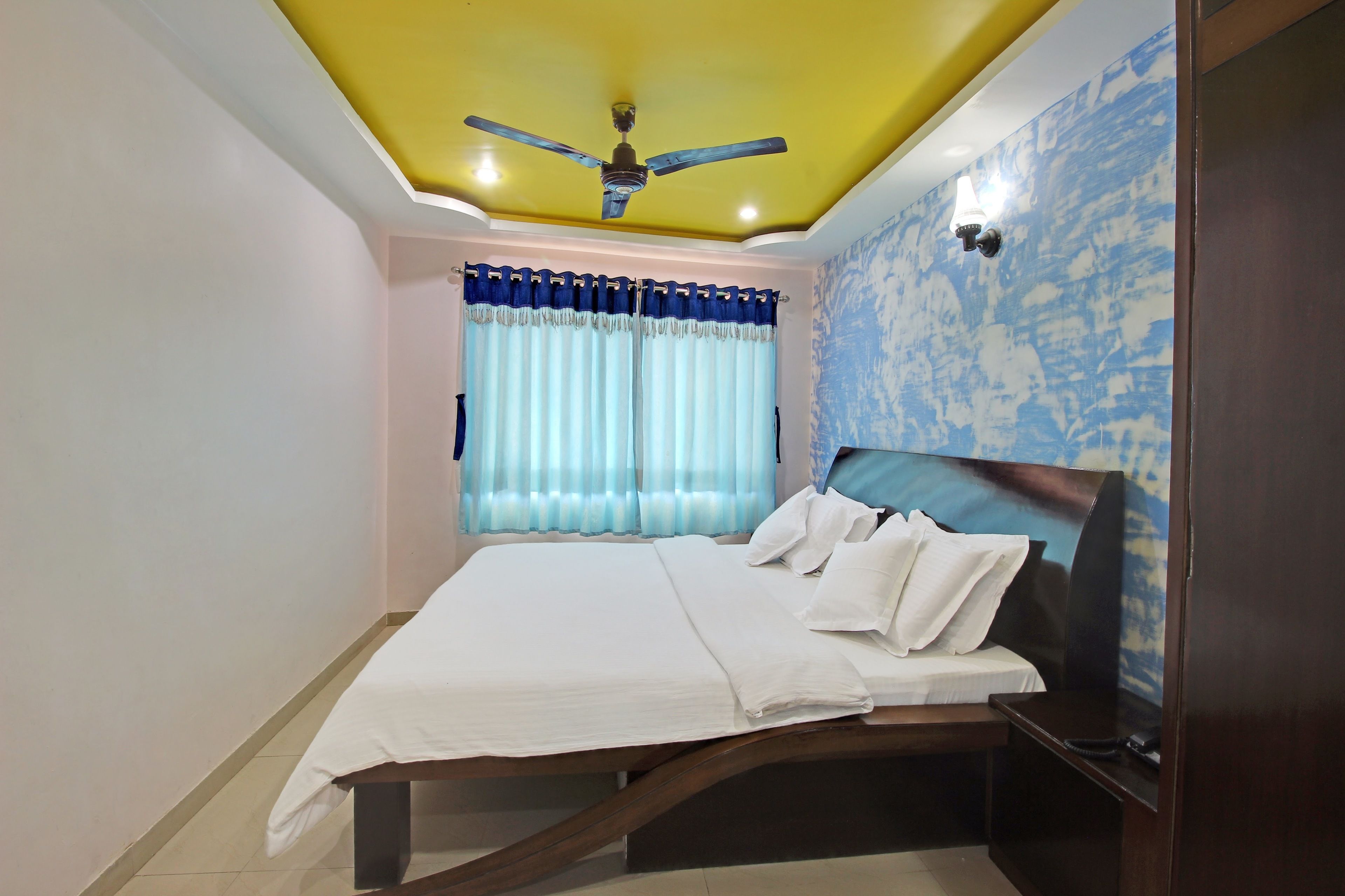 A bedroom with a bed, a ceiling fan, a window with curtains, and a wall with a blue pattern.