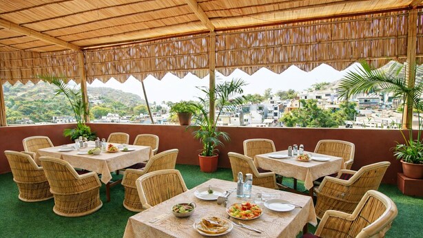 Un área de comedor al aire libre con mesas y sillas de mimbre, rodeada de plantas en macetas y vistas a un paisaje urbano.