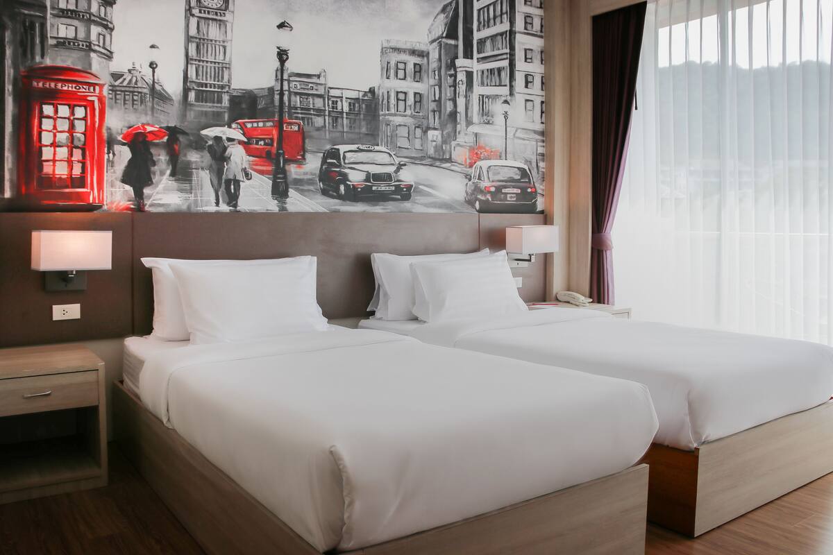 A hotel room with two beds, a large cityscape mural on the wall, and a window with curtains.