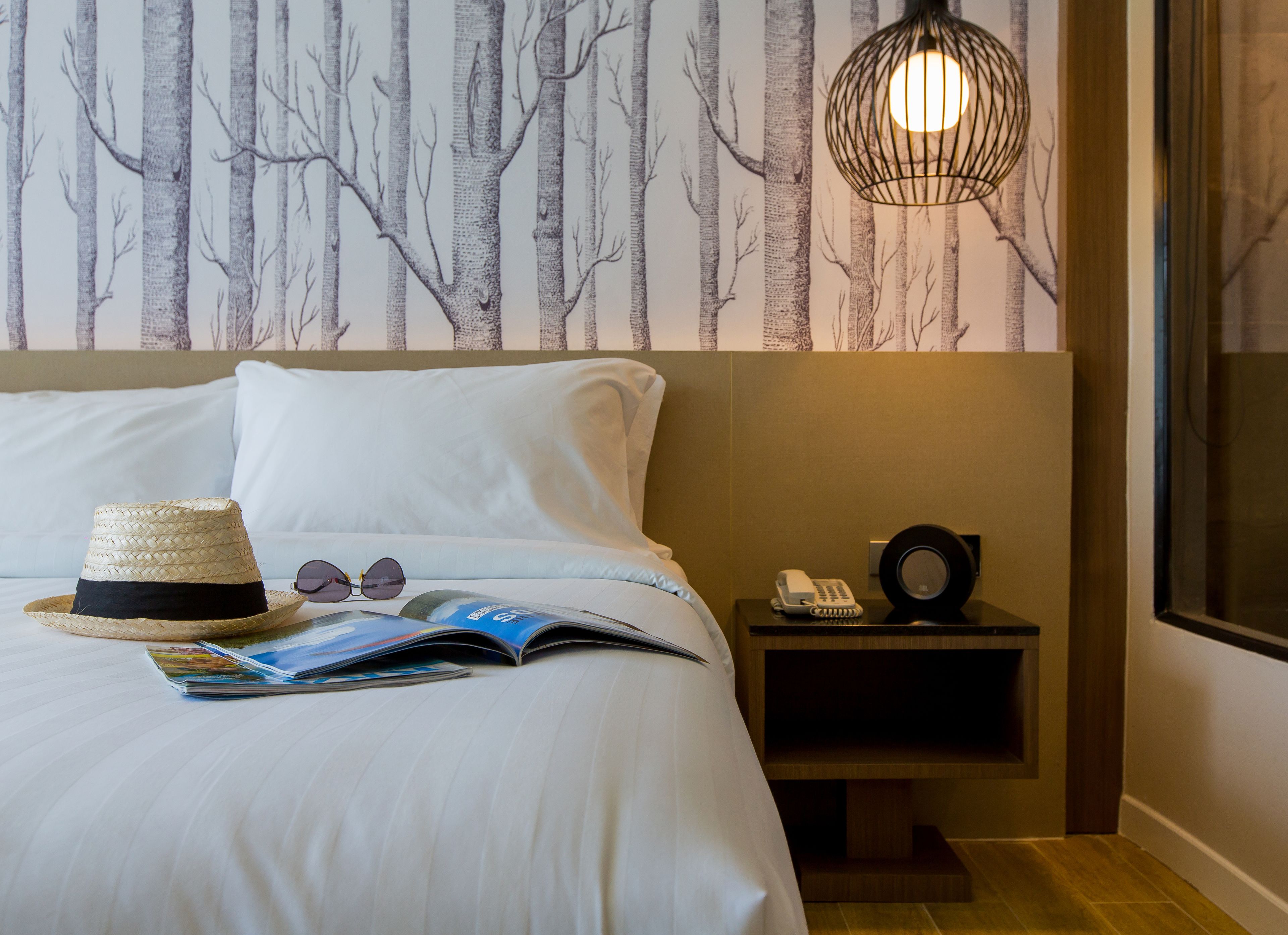 A neatly made bed with a magazine, a hat, and sunglasses on it. There is a bedside table with a phone and a lamp. The wall has a tree pattern wallpaper.