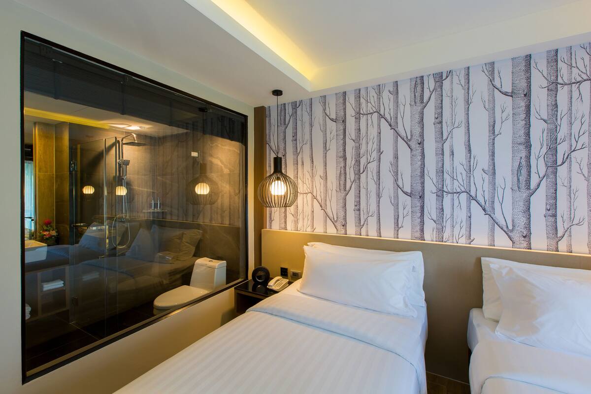 A modern hotel room with a large mirror, a bed with white linens, a bedside table with a lamp, and a wall with a tree pattern wallpaper.