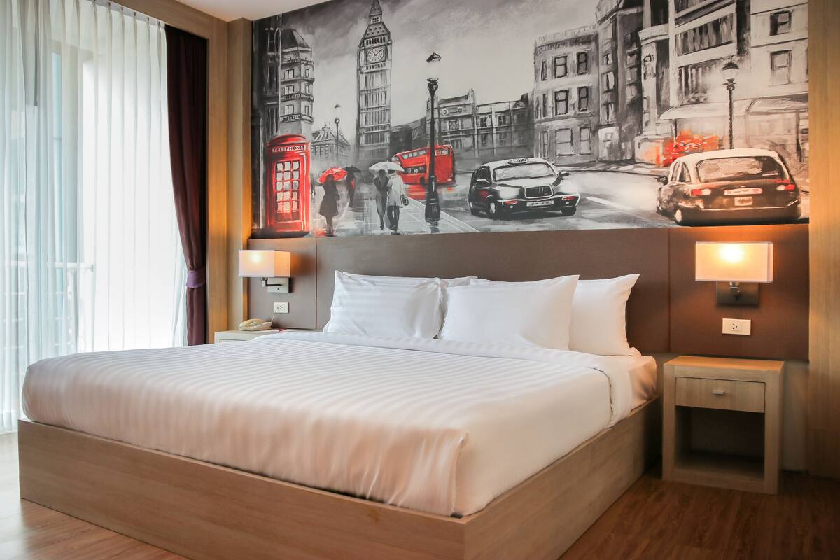 A hotel room with a large bed, a nightstand with a lamp, and a wall mural of a city scene.