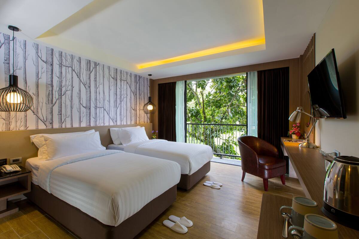A hotel room with two beds, a desk with a coffee maker, a TV, and a balcony with a view of trees.