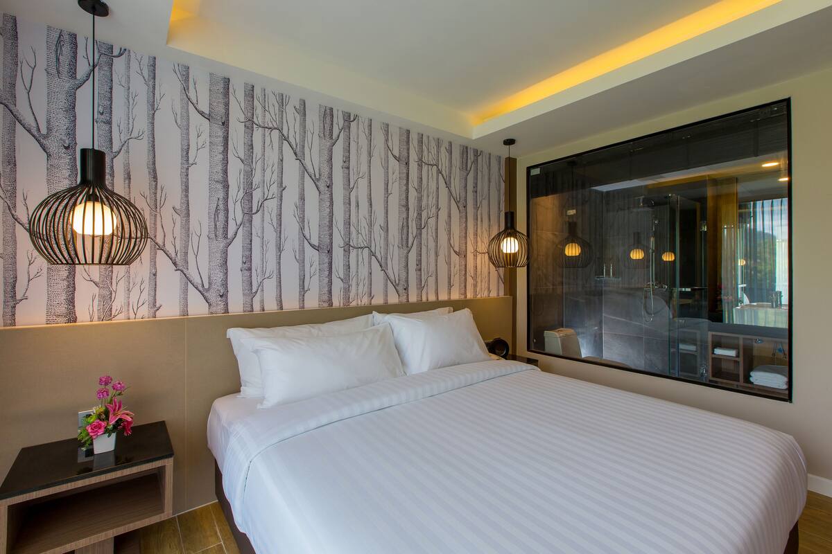 A hotel room with a large bed, a nightstand with a vase of flowers, a wall with a tree pattern, and a large mirror reflecting the room.