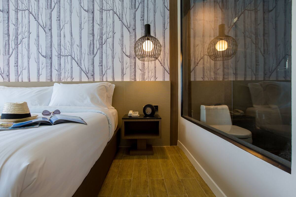 A hotel room with a bed, a nightstand with a phone and a book, a window with a view of a toilet, and a wall with a tree pattern.