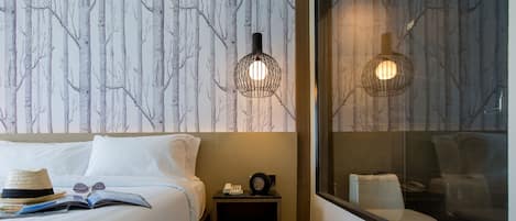Habitación de hotel con una cama, una mesita de noche con un teléfono y un libro, una ventana con vista a un inodoro y una pared con un diseño de árbol.