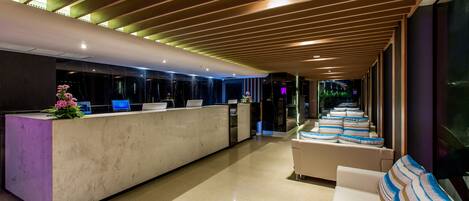 A modern hotel lobby with a reception desk, seating area, and a long corridor with windows.