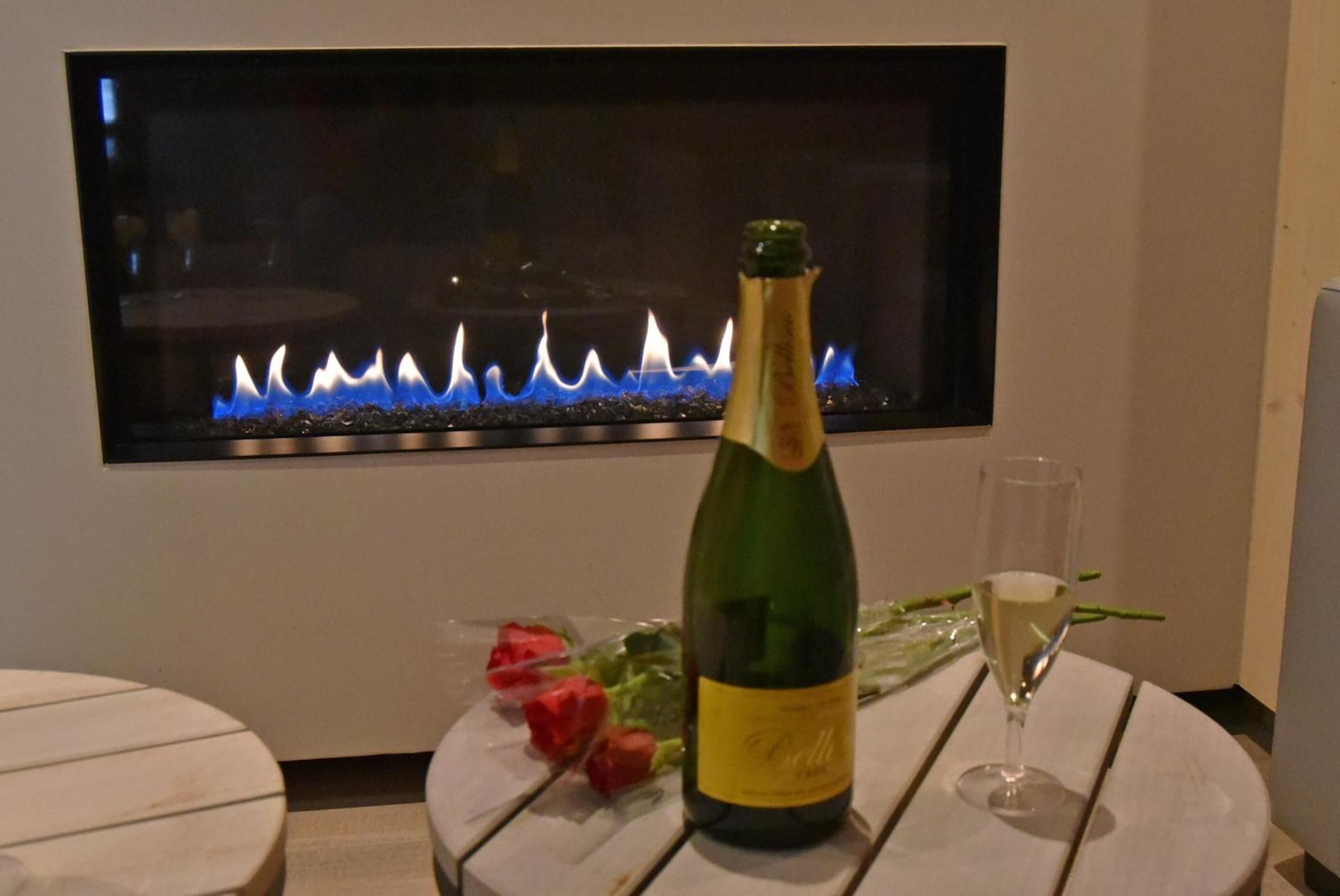 A bottle of champagne, a glass, and a bouquet of roses on a table with a modern fireplace in the background.