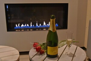 Eine Champagnerflasche, ein Glas und ein Rosenstrauß auf einem Tisch, im Hintergrund ein moderner Kamin.