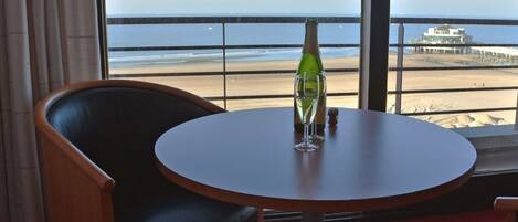 A room with a round table, a chair, and a view of the beach and ocean through large windows.