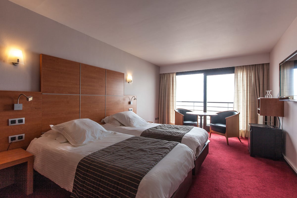 A hotel room with two beds, a desk, a chair, a TV, and a large window with curtains.