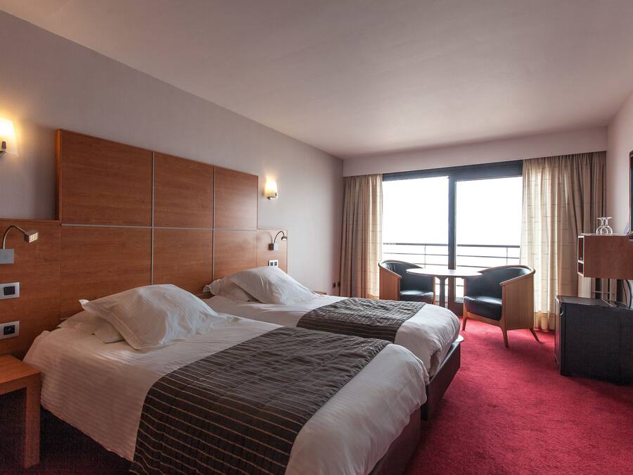 Une chambre d’hôtel avec deux lits, un bureau, une chaise, une télévision et une grande fenêtre avec des rideaux.