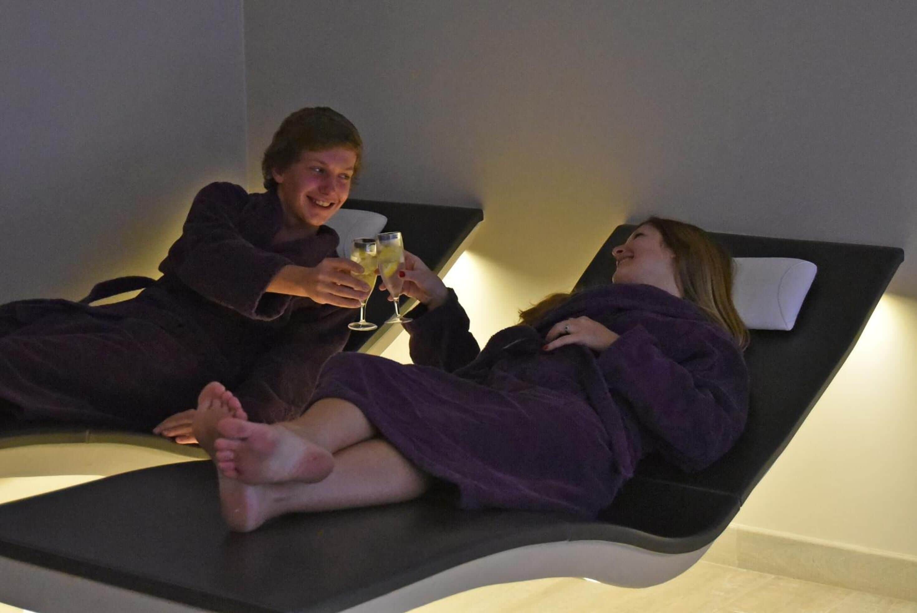 Two people in purple robes on a reclining chair with a massage table, holding glasses.