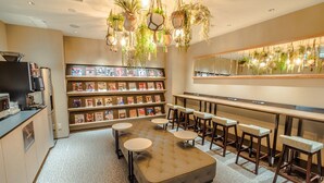 A cozy reading area with a tufted ottoman, round tables, and bar stools. A bookshelf filled with books is visible in the background. - Glansit Akihabara Comfort Capsule Hotel (Tokyo)