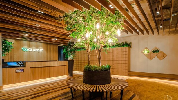 A reception area with a wooden reception desk, a large potted tree, and a modern design with wooden paneling and recessed lighting. - Glansit Akihabara Comfort Capsule Hotel (Tokyo)