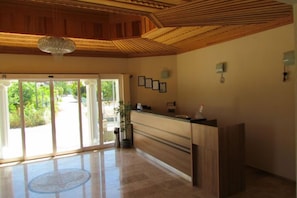 A reception area with a wooden reception desk, a chandelier, framed pictures on the wall, and a view of greenery outside.
