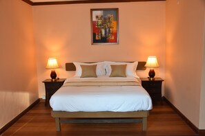 Una cama bien hecha con ropa de cama blanca y marrón, flanqueada por dos mesitas de noche con lámparas, en una habitación con suelo de madera y un cuadro en la pared.