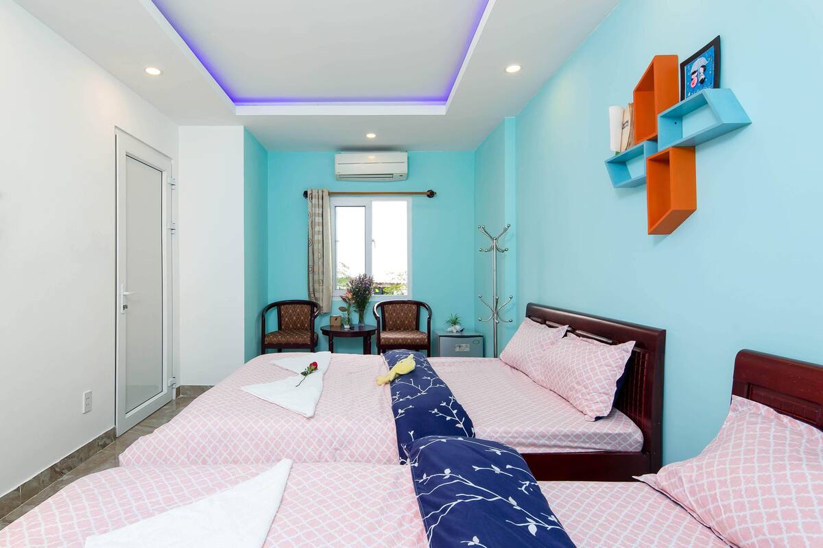 Une chambre d’hôtel avec deux lits, une fenêtre avec des rideaux, une petite table avec une plante et une étagère fixée au mur, sur laquelle sont posés divers objets.