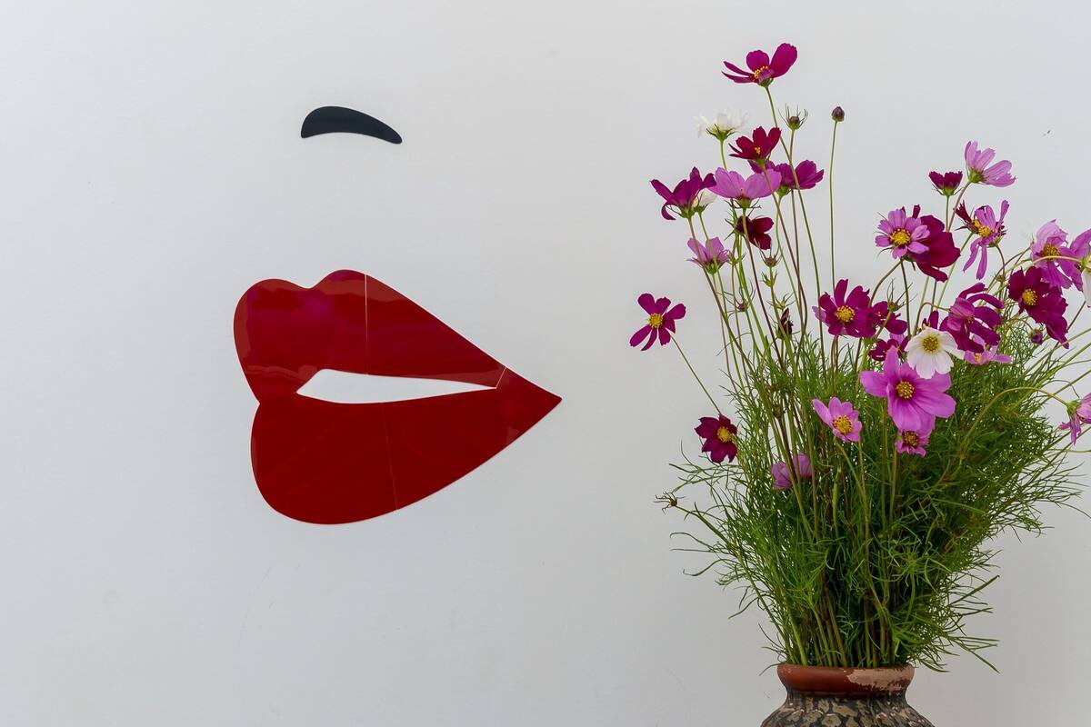 Un mur orné d’un motif de visage peint à la pochoir, un vase rempli de fleurs et des pommes posées sur une table.