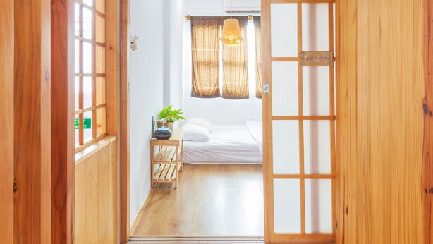Une pièce de style japonais traditionnel, avec une porte coulissante en bois, un lit recouvert de draps blancs, une petite table de chevet ornée d’une plante et un luminaire suspendu.