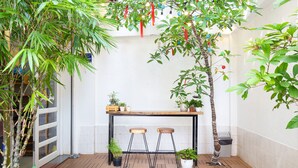 Un petit espace extérieur avec une table en bois et deux tabourets, entouré de plantes en pot et d’un arbre en bambou.