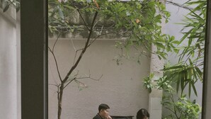 Deux personnes sont assises à une table dans un espace extérieur doté d’un sol carrelé, entourées de verdure et d’un mur sobre.