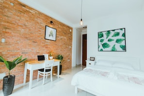 Ein modernes Zimmer mit Backsteinwand, einem weißen Schreibtisch mit Laptop, einem Bett mit weißer Bettwäsche und zwei Zimmerpflanzen.