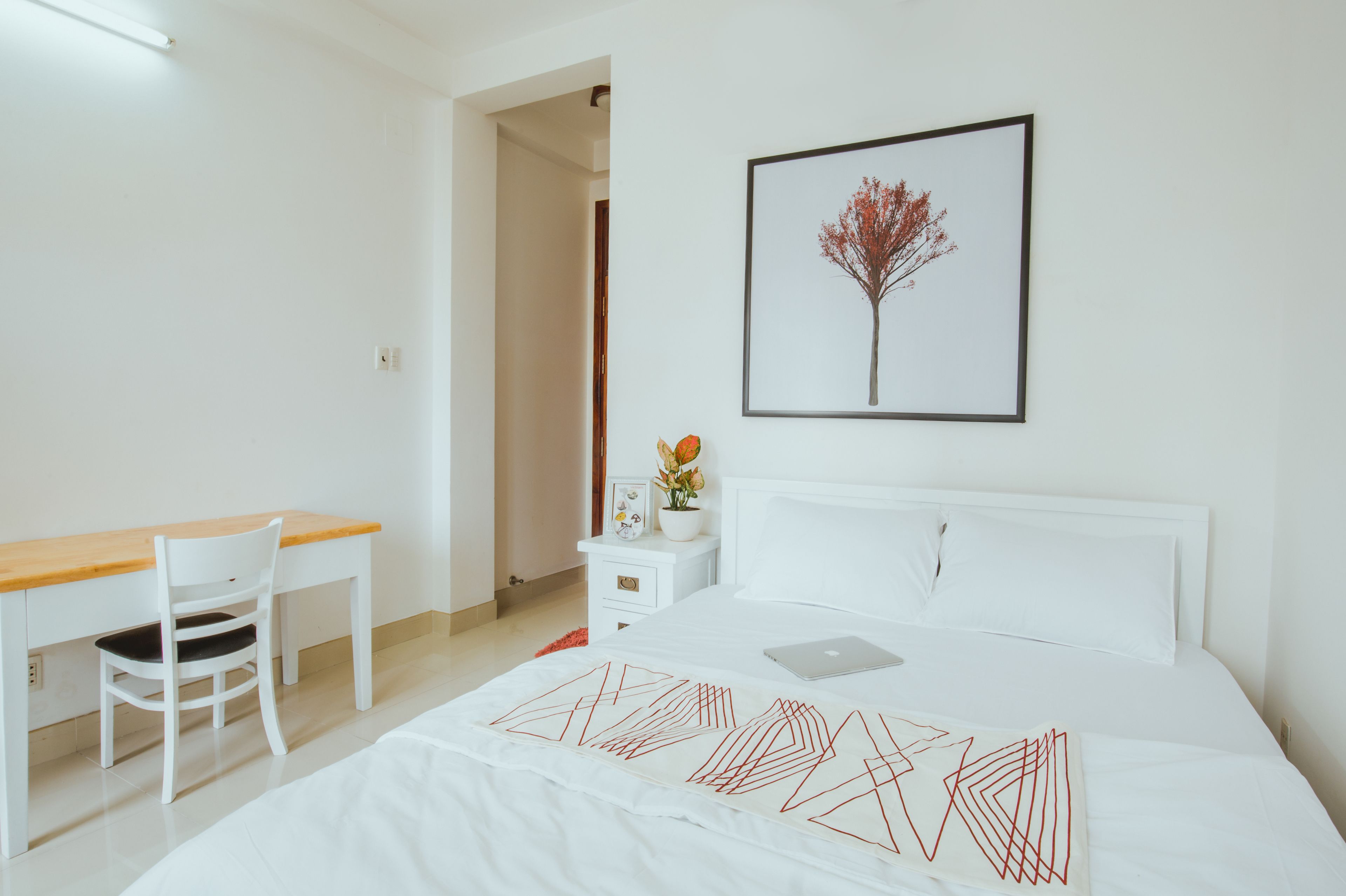 A white bedroom with a bed, a desk, and a chair. There is a framed picture on the wall and a potted plant on the nightstand.
