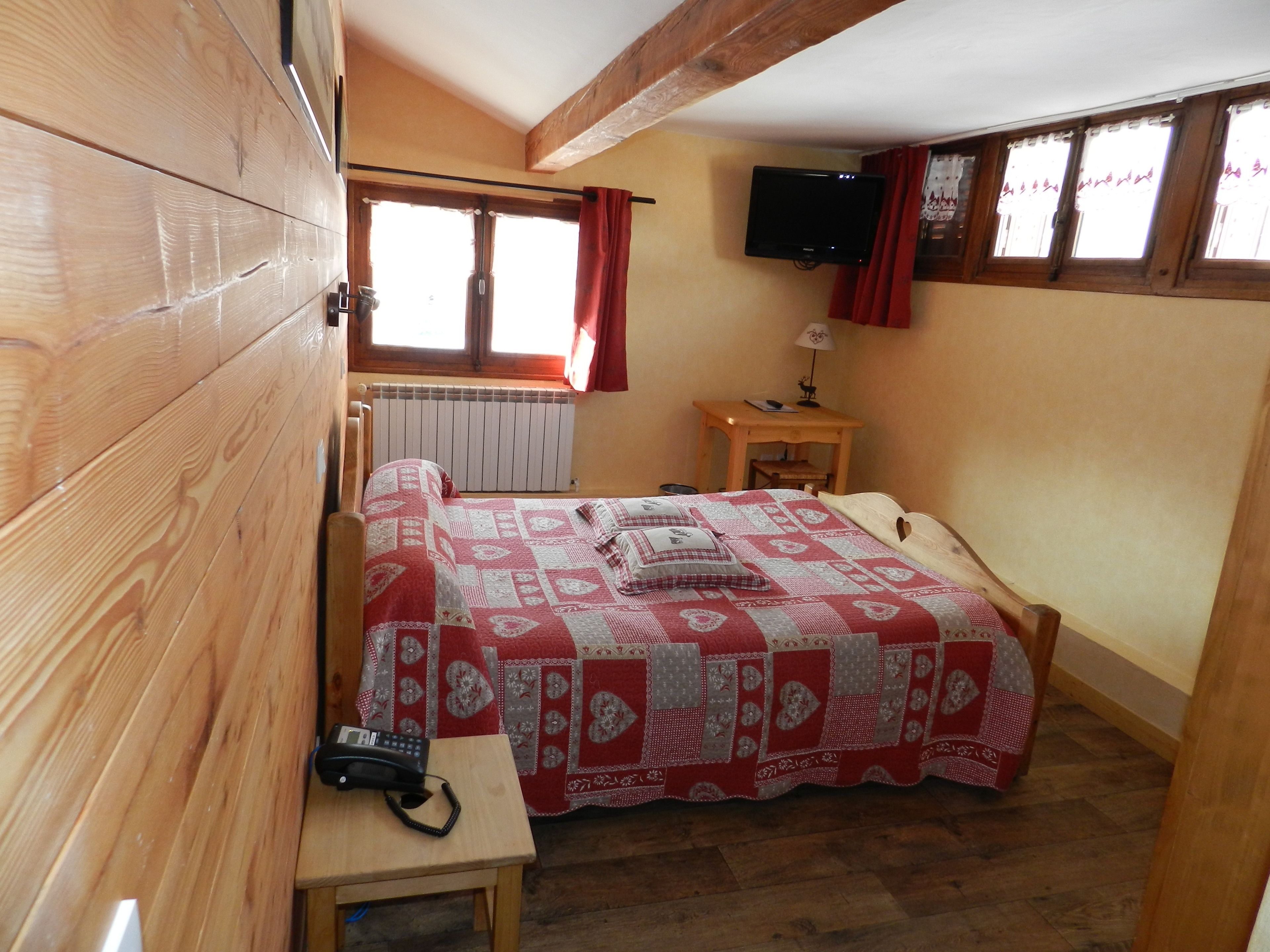 Una camera da letto con un letto di legno, un comodino con un telefono, una finestra con tende, un termosifone e una TV appesa al muro.