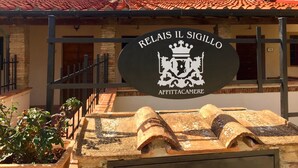Une enseigne pour le « RELAIS IL SIGILLO AFFITTACAMERE », avec un emblème en forme de blason, un toit en tuiles et un escalier menant à l’entrée du bâtiment.