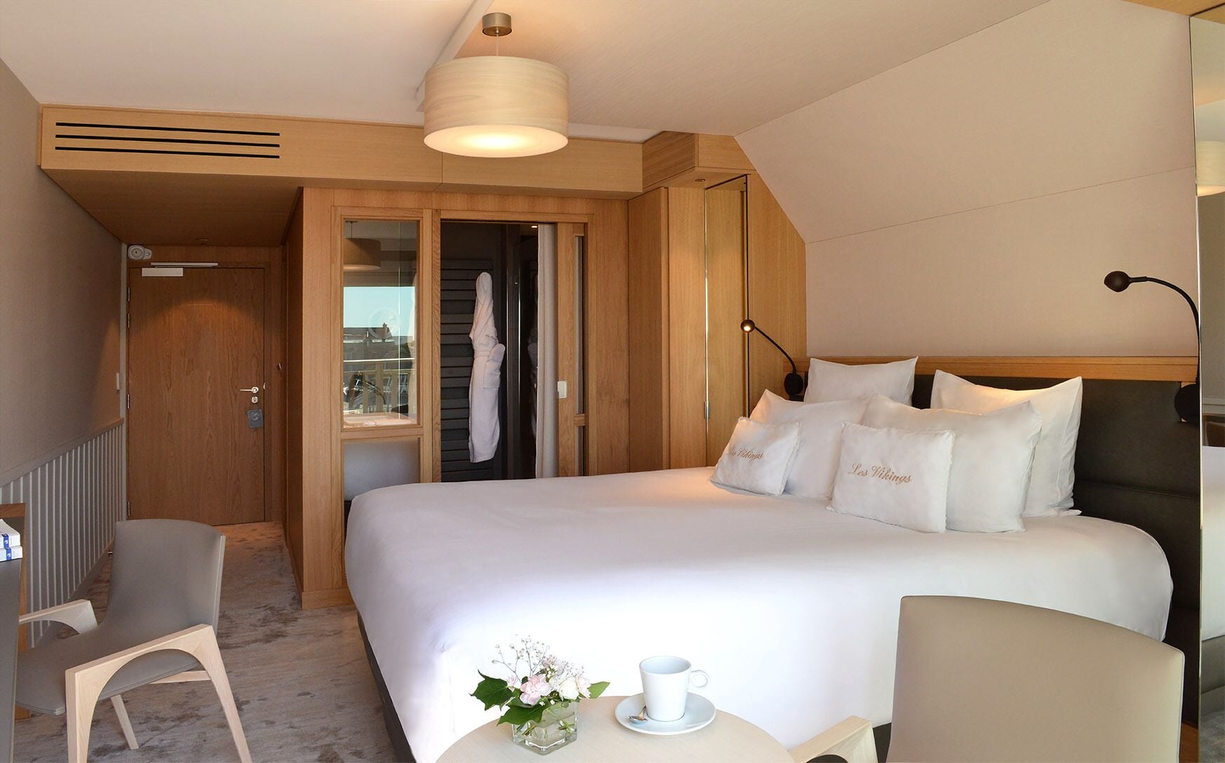 Habitación de hotel con una cama grande, un escritorio con silla, una mesita con una taza y flores, y un armario con una bata colgada.