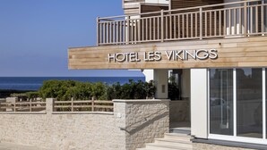 Hotelli nimeltä ”HOTEL LES VIKINGS”, jossa on puinen julkisivu, kiviseinä ja näkymä merelle.