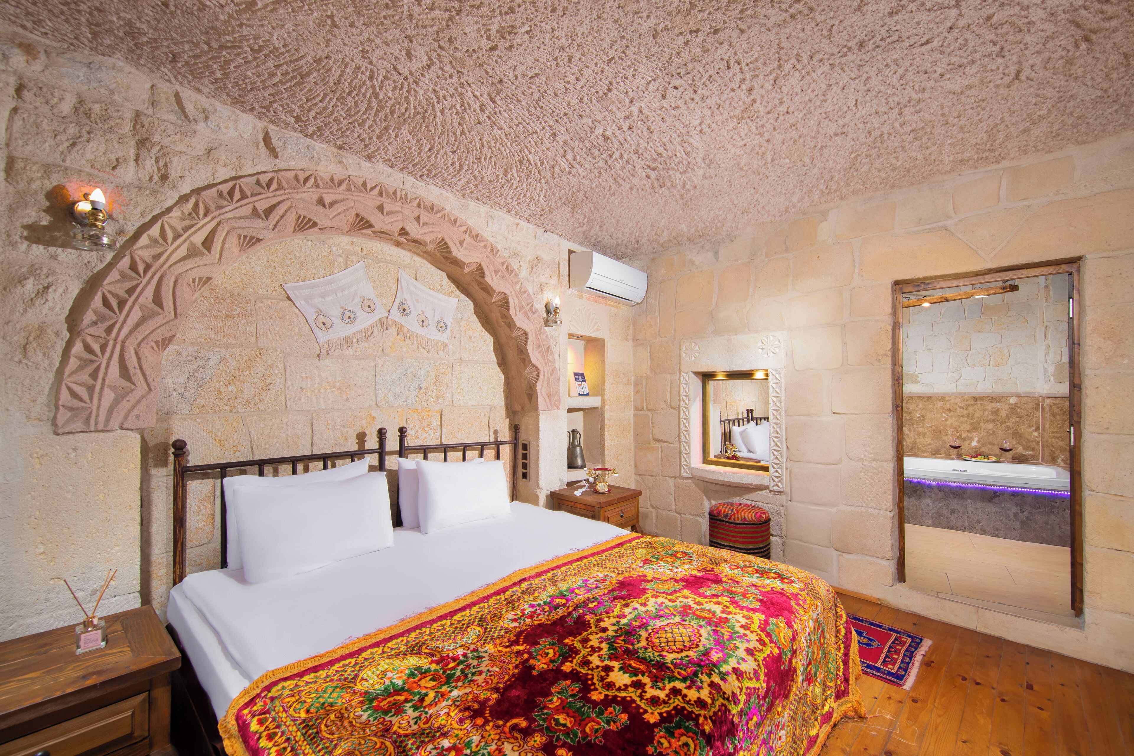 Un dormitorio con pared de piedra, una cama con ropa de cama blanca, una manta colorida, una mesita de noche, una lámpara y una ventana con vista a un baño.