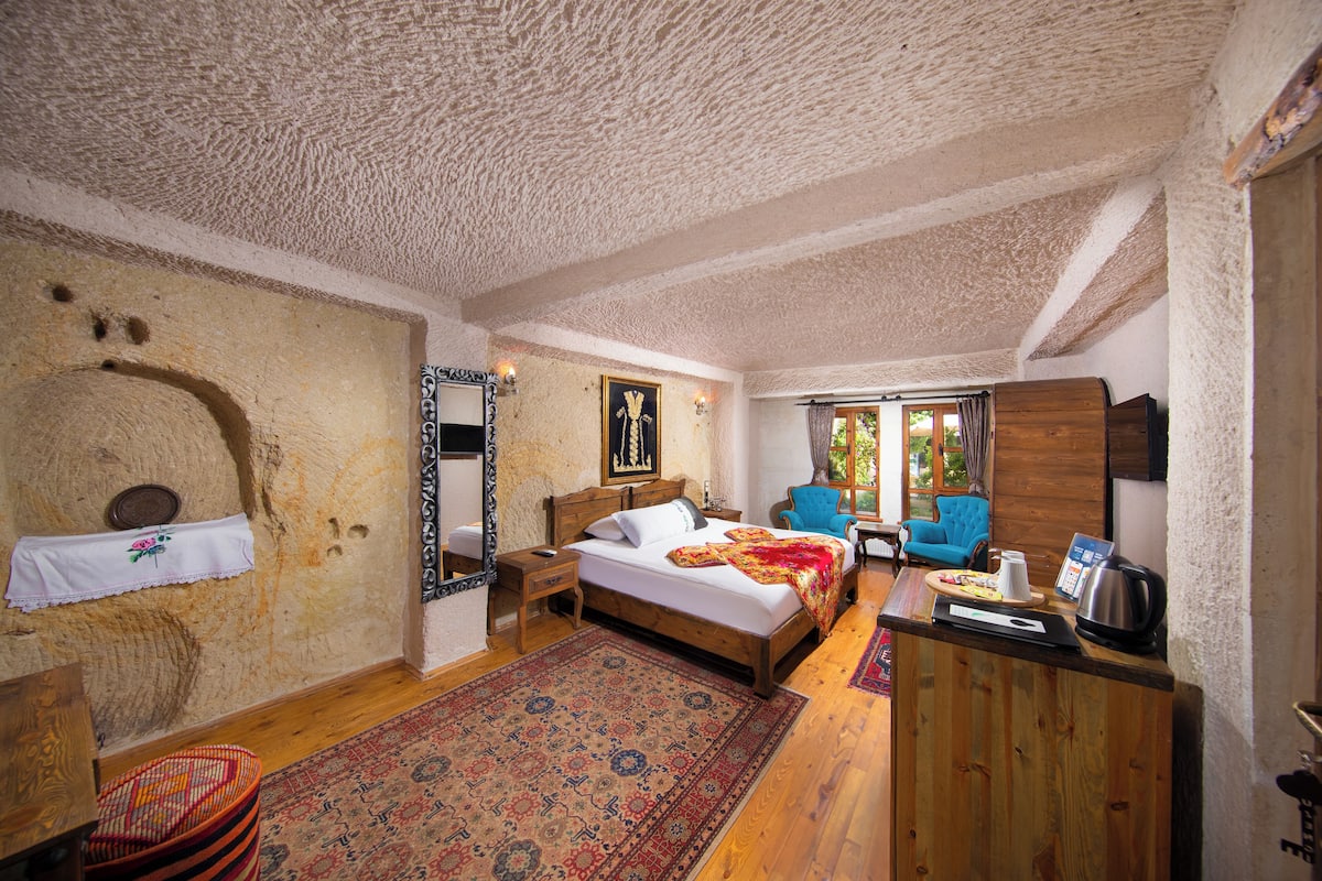 Un dormitorio con una cama de madera, una mesita de noche, un sillón azul, un escritorio de madera con una tetera y una alfombra estampada.