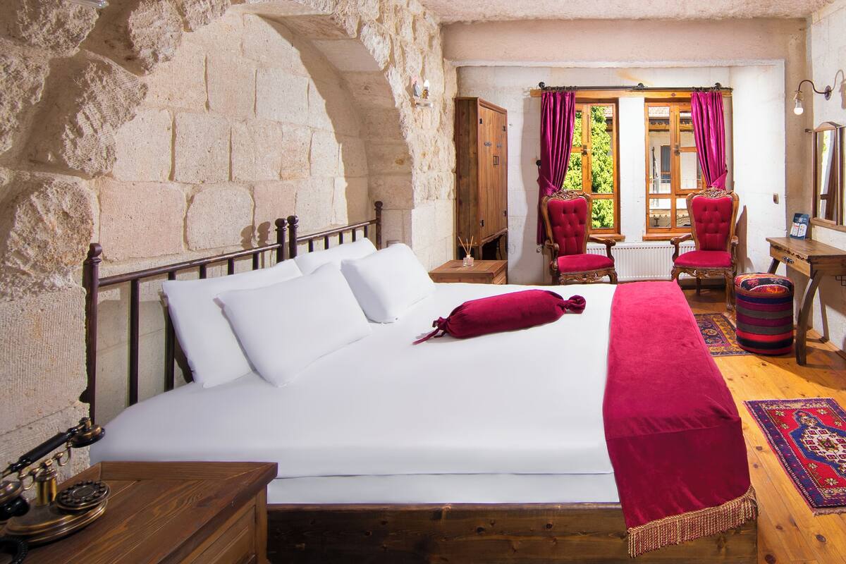 Un dormitorio con pared de piedra, una cama de madera, una silla roja, un escritorio de madera y una ventana con cortinas.