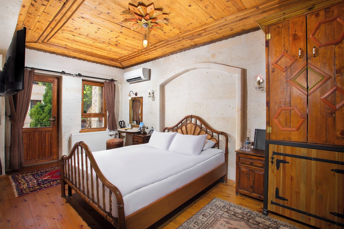 Una cama con estructura de madera y ropa de cama blanca, un armario de madera, un escritorio con una silla y un televisor en la pared.