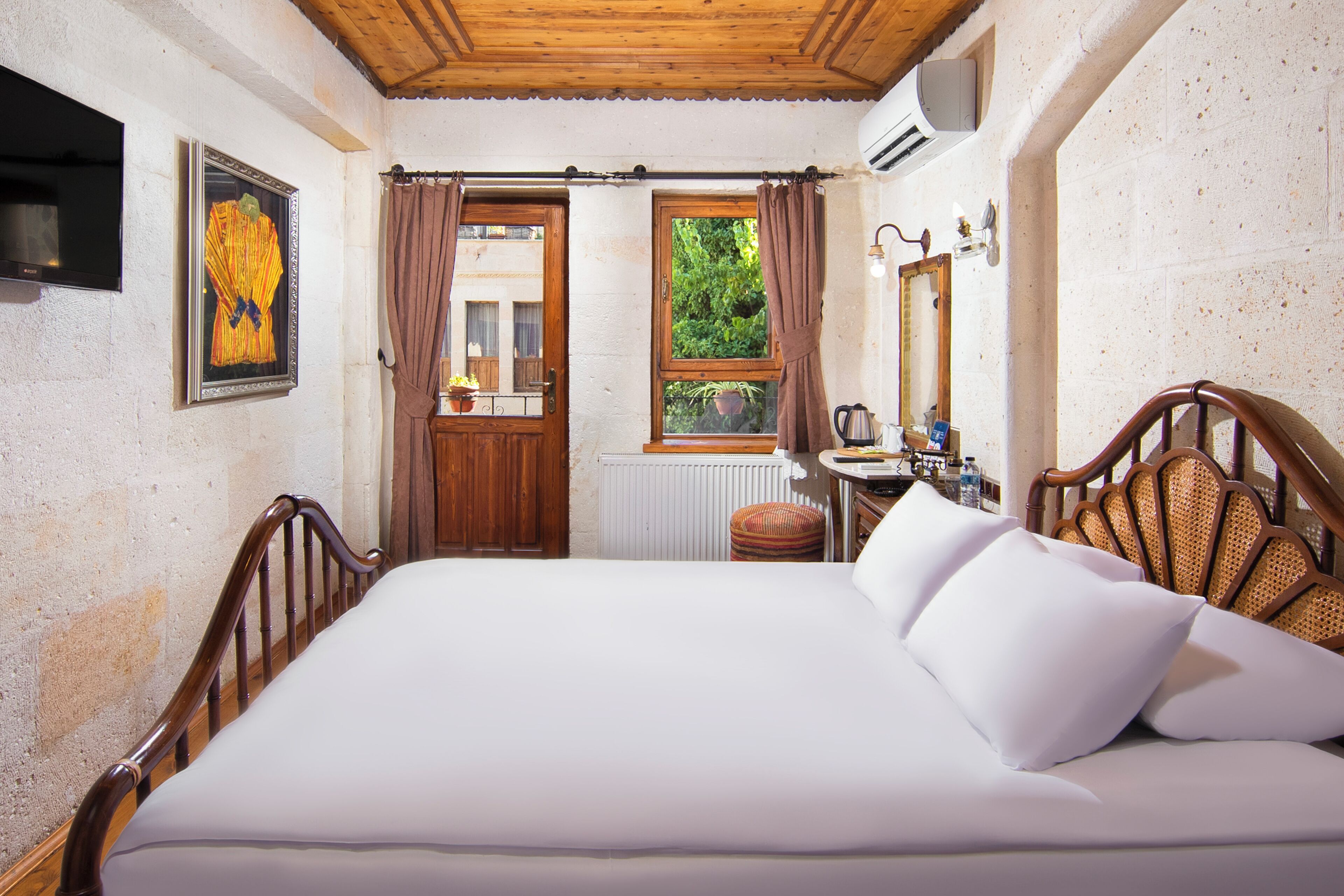 Un dormitorio con una cama grande, cabecera de madera, una mesita de noche con lámpara, una ventana con cortinas y un cuadro en la pared.