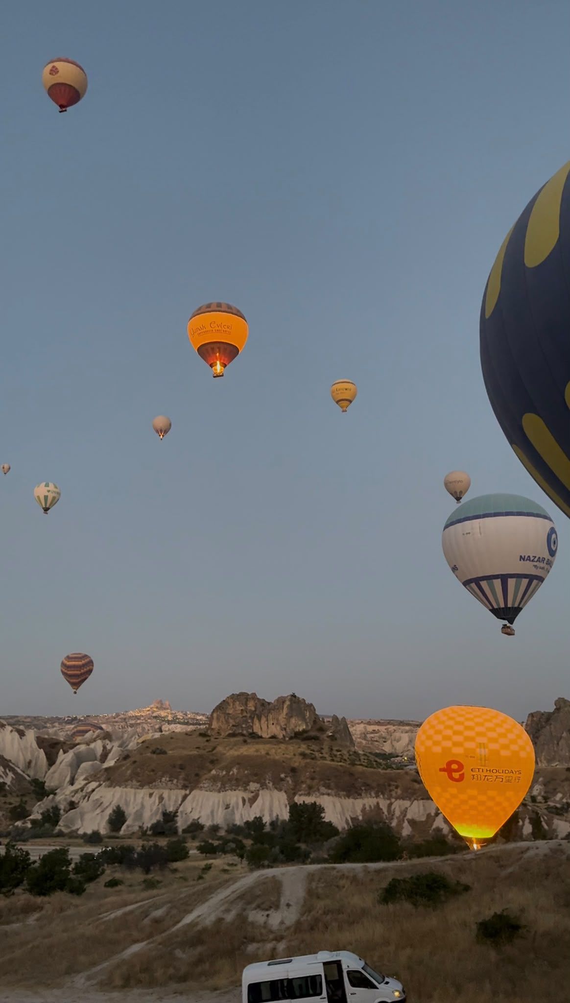 Un paisaje con varios globos aerostáticos en el cielo y un vehículo blanco en el suelo.