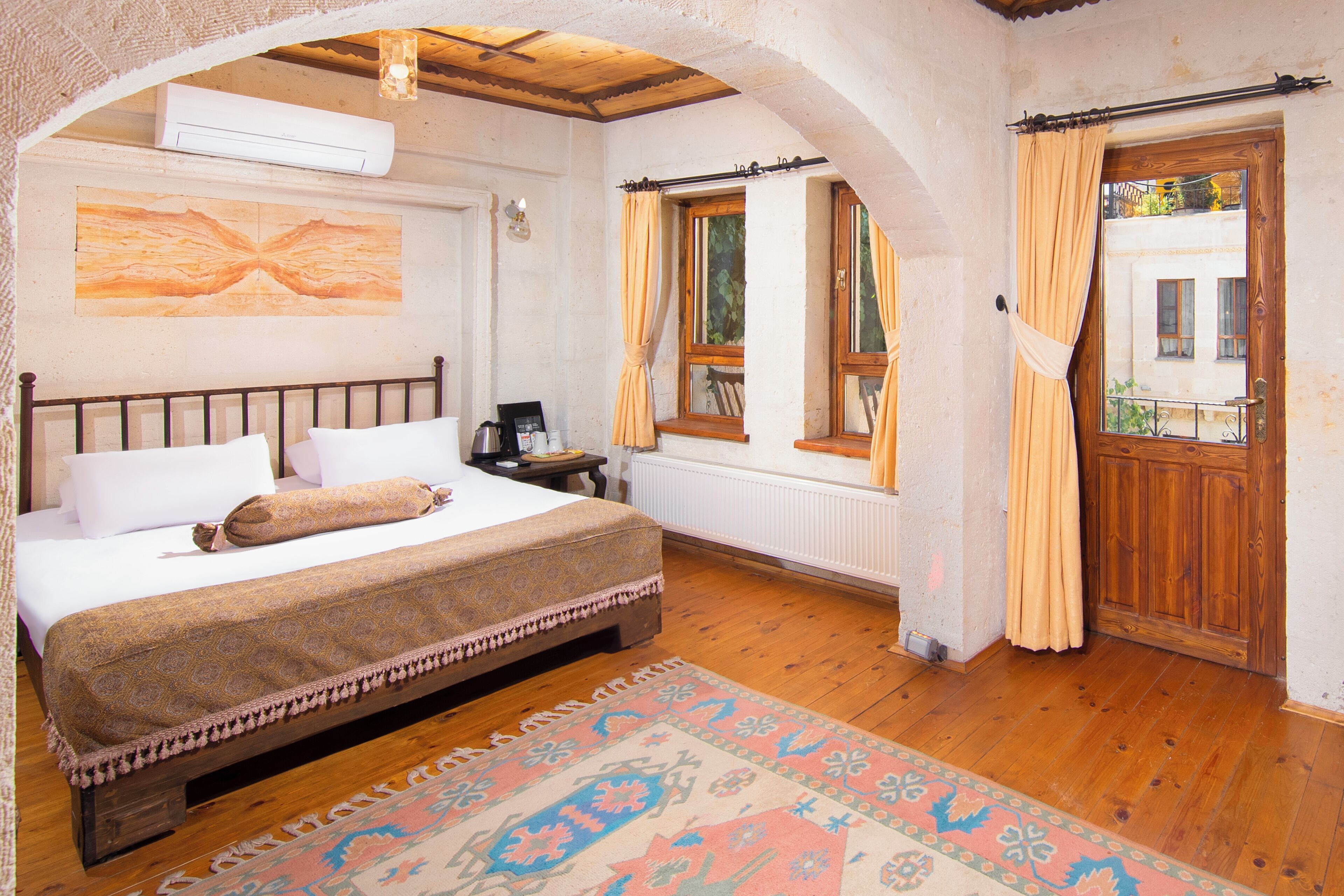 Un dormitorio con una cama grande, suelo de madera, alfombra estampada y una ventana con cortinas.