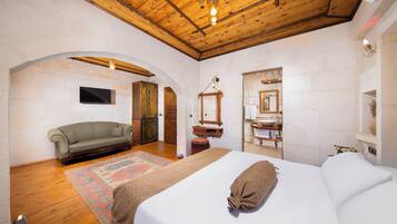 Un dormitorio con una cama grande, techo de madera, televisor y un baño visible a través de un arco.