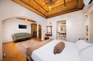 Un dormitorio con una cama grande, techo de madera, televisor y un baño visible a través de un arco.