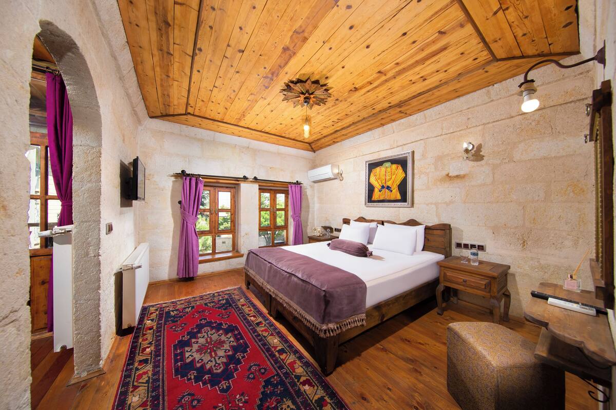 Un dormitorio con techo de madera, una cama grande, una mesita de noche, una silla y una alfombra roja estampada.