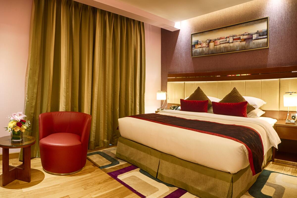 Ein Hotelzimmer mit einem großen Bett, einem roten Sessel, einem kleinen Tisch mit Blumenvase und einem Gemälde an der Wand.