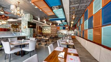 A restaurant with a long counter, multiple tables, and chairs. There are ceiling fans and a TV mounted on the wall.