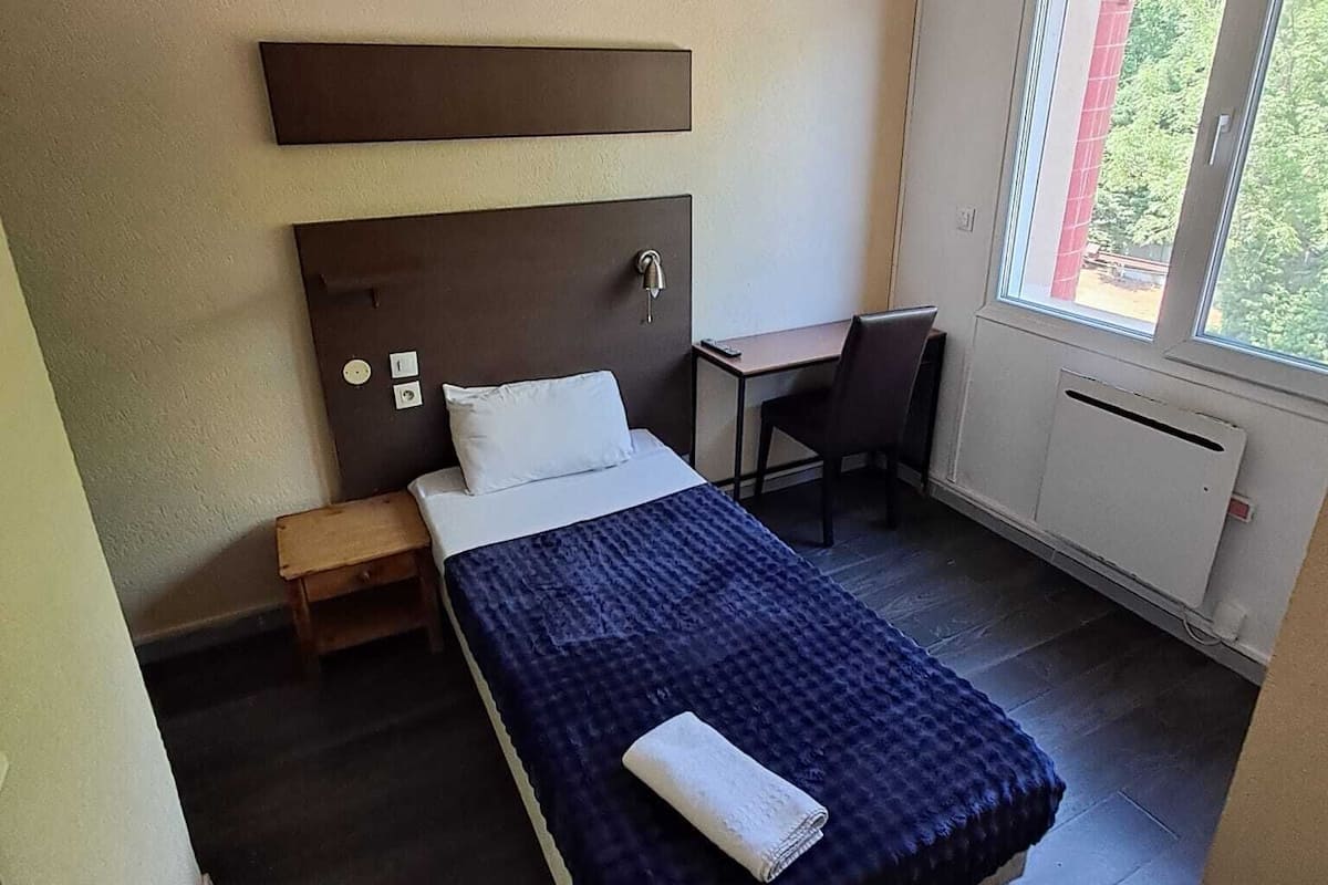 A hotel room with a bed, a desk, a chair, a small table, and a window with a view of greenery.