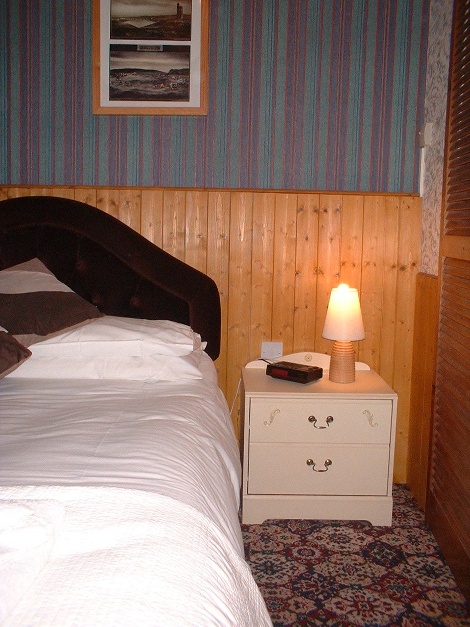 A bed with a wooden headboard, a nightstand with a lamp, and a framed picture on the wall.