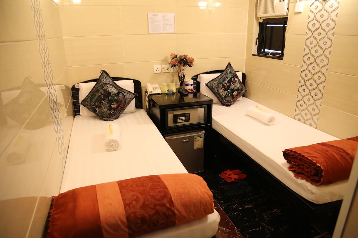 A hotel room with two single beds, a small table with a phone and a vase of flowers, and a window with a patterned curtain.