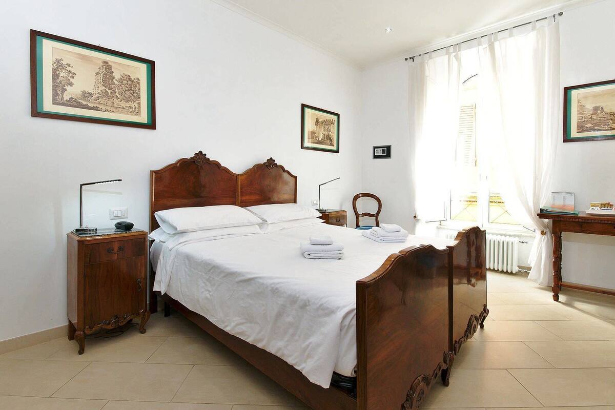 Una cama bien hecha con sábanas blancas, cabecera de madera y mesita de noche con lámpara.